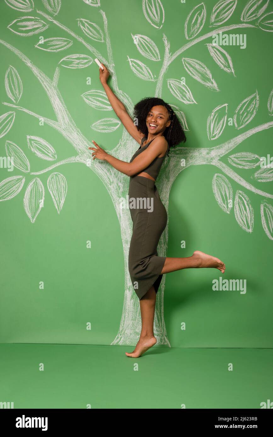 Lächelnde junge Frau, die auf einem Bein steht und einen Baum mit Kreide auf grünem Hintergrund zeichnet Stockfoto