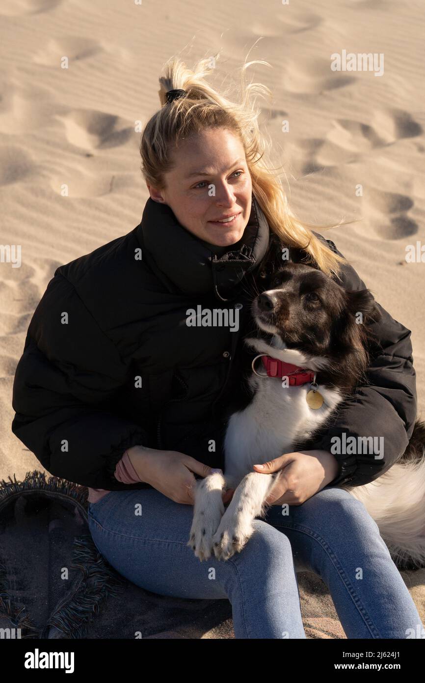 Lächelnde junge blonde Frau, die mit Hund auf Sand sitzt Stockfoto