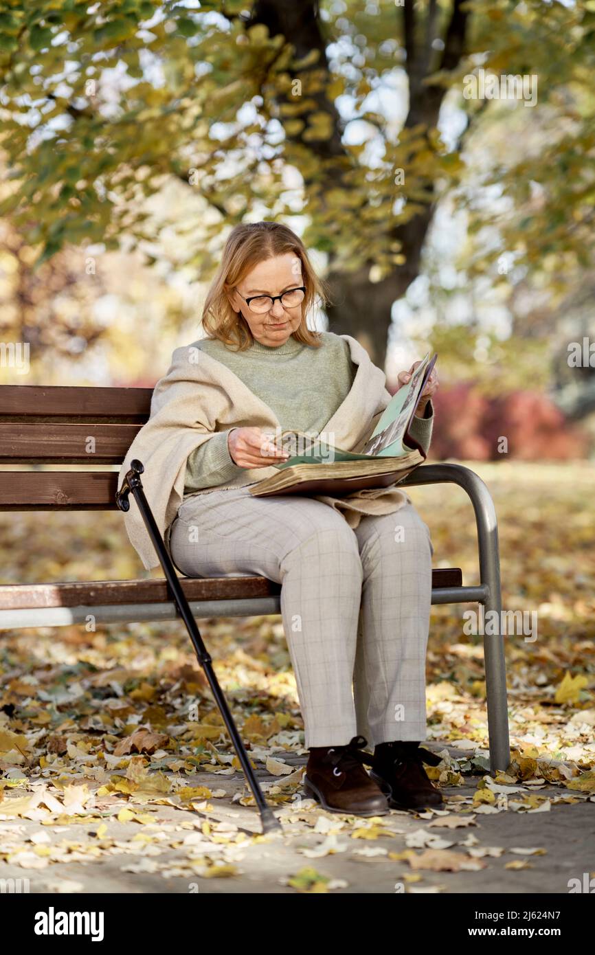 Ältere Frau, die das Fotoalbum anschaut, sitzt mit Gehstock auf der Bank im Park Stockfoto