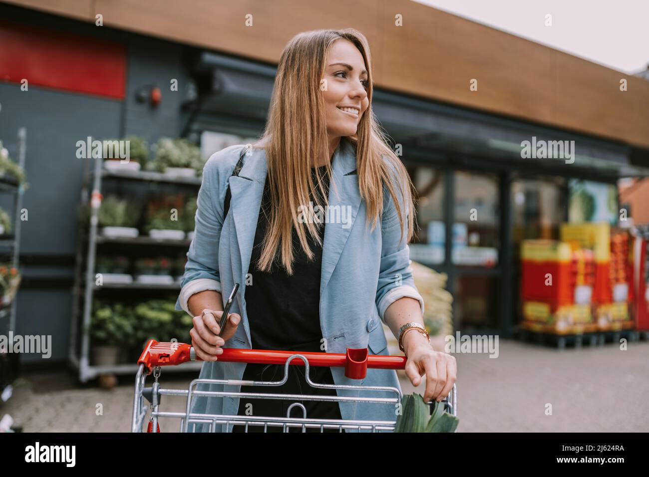 Lächelnde Frau mit Smartphone und Einkaufswagen, die vor dem Geschäft steht Stockfoto