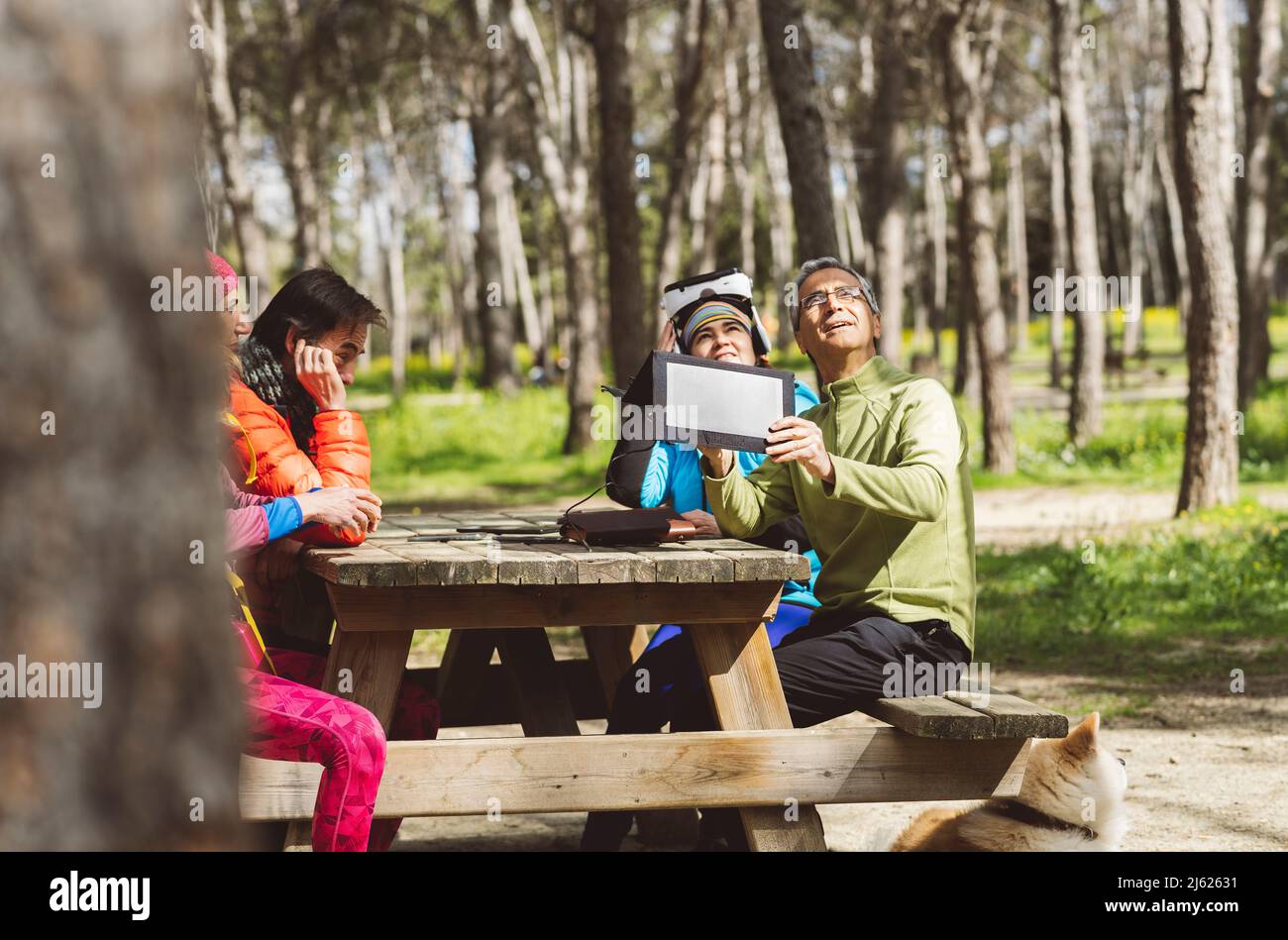 Reifer Mann, der Solarpanel unter Sonnenlicht anpasst, sitzt mit Freunden am Picknicktisch im Wald Stockfoto
