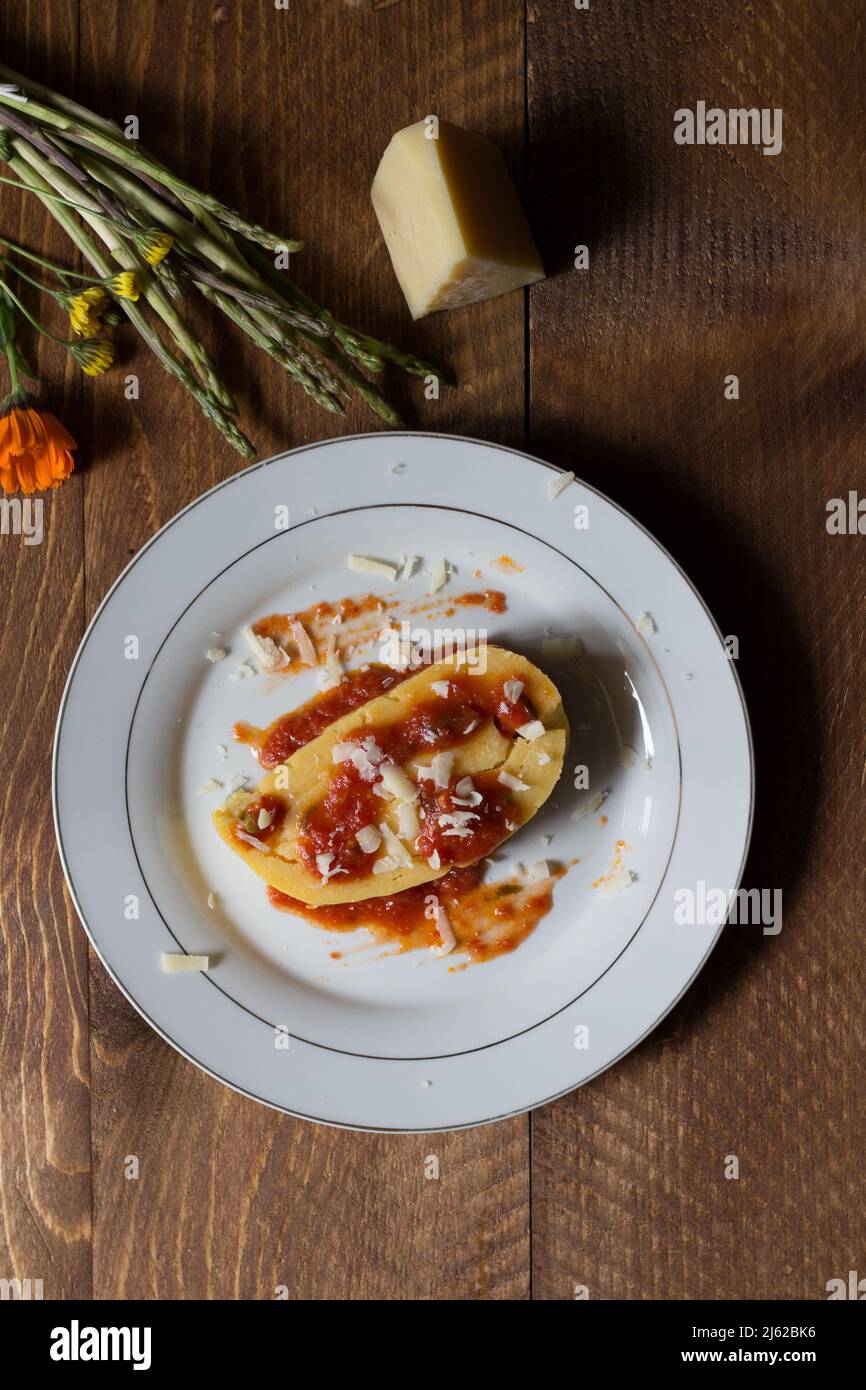 Mais-Polenta gerollt mit Spargel und gekrönt mit Soße und Käse Flocken auf weißem Teller und dunklem Holz Hintergrund Stockfoto