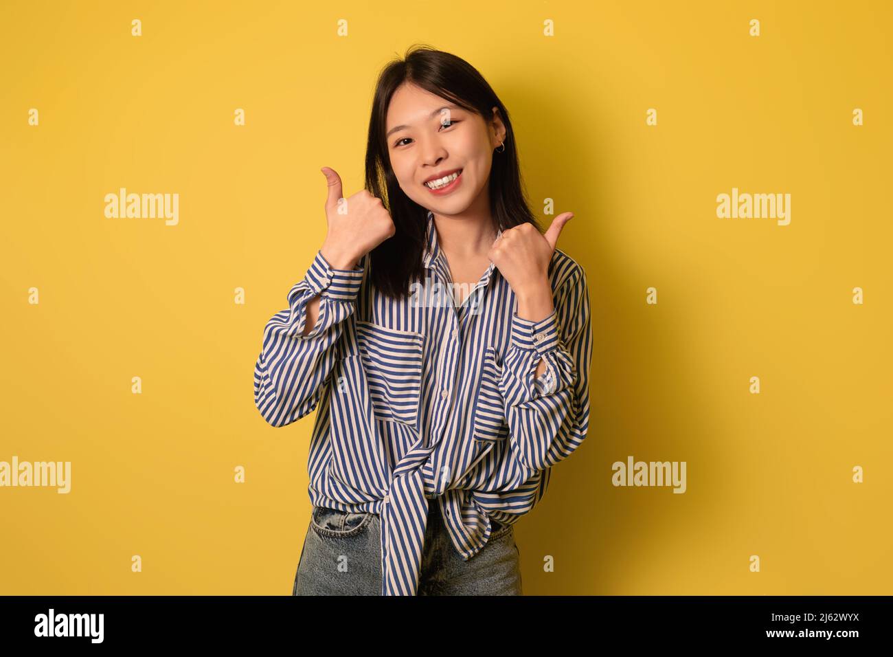 Lächelnde junge asiatische Frau, die mit beiden Händen Daumen nach oben zeigt und etwas auf gelbem Studiohintergrund empfiehlt Stockfoto