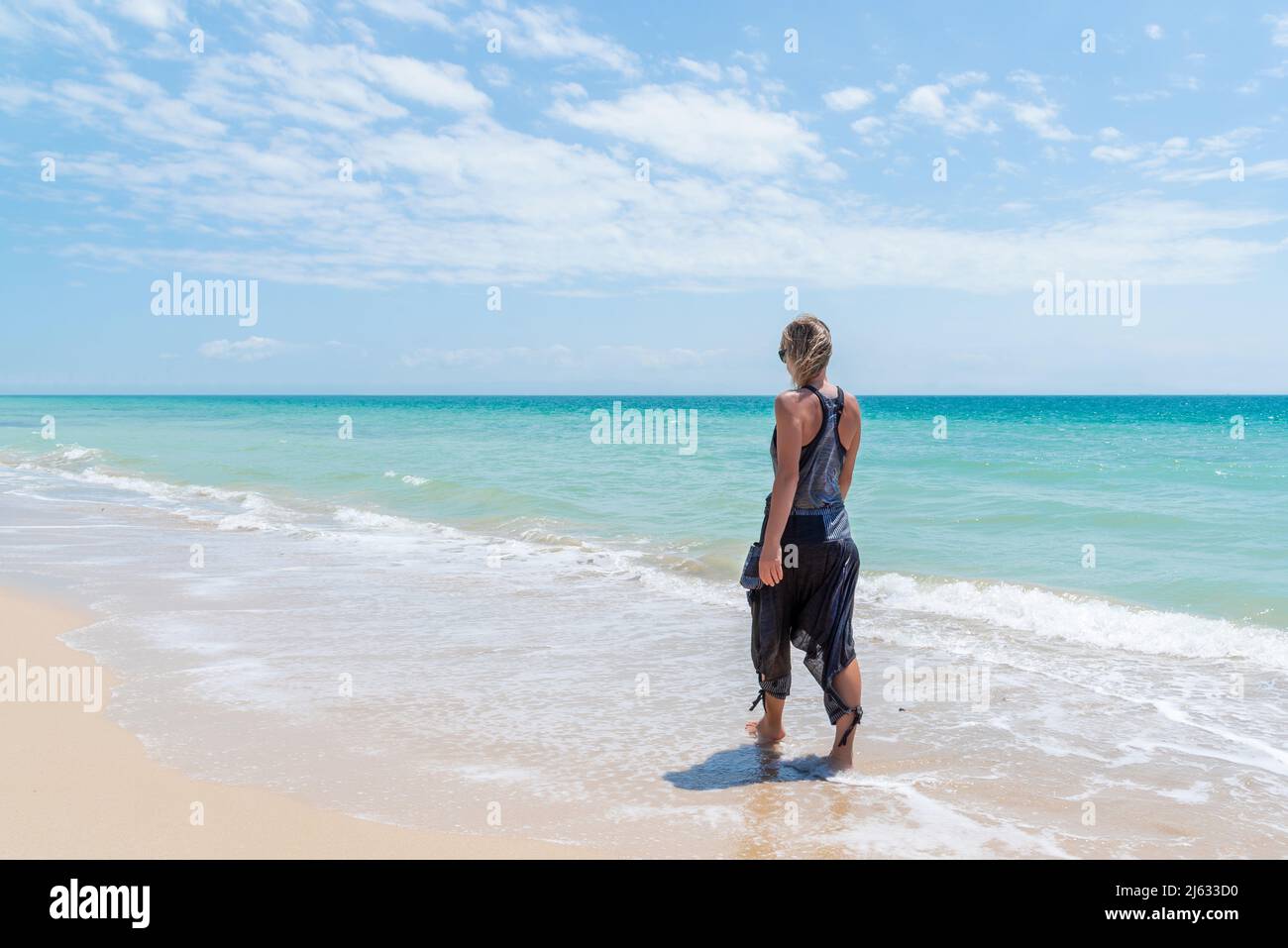 Kaukasische blonde Frau, die im Urlaub in legerer Kleidung am Ufer eines goldenen Sandstrands entlang geht Stockfoto