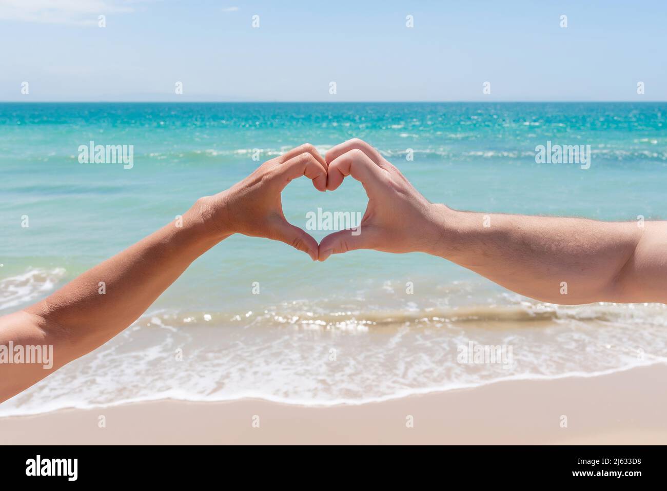 Hände bilden ein Herz vor einem Strand aus goldenem Sand und türkisfarbenem Wasser. Liebe im Paradies Stockfoto