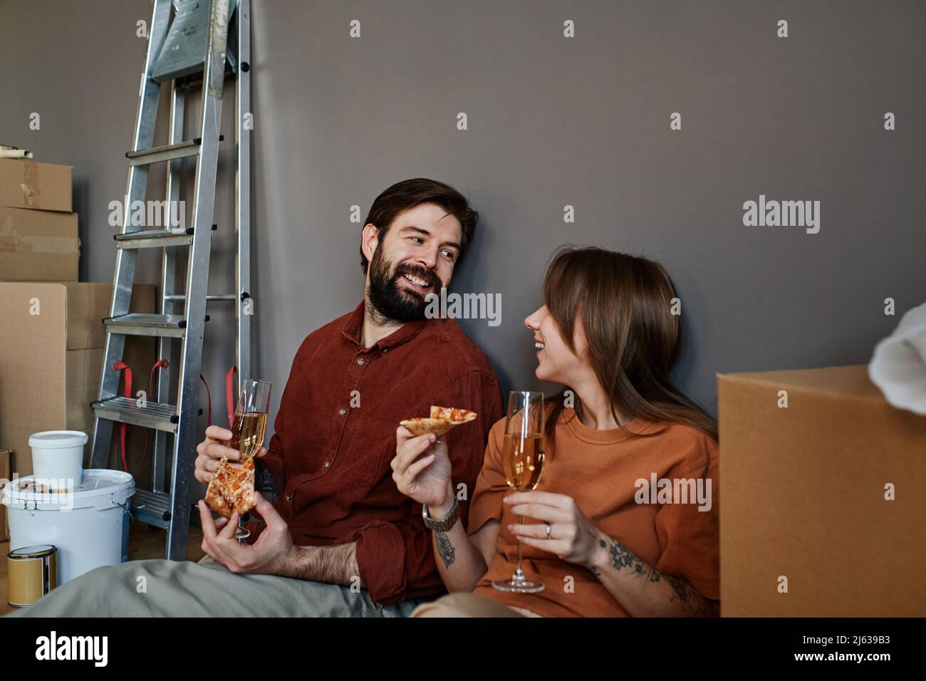 Horizontale Aufnahme eines verliebten jungen Mannes und einer verliebten Frau, die auf dem Boden sitzen, Pizza essen und Sekt trinken, um den Umzug in eine neue Wohnung zu feiern Stockfoto