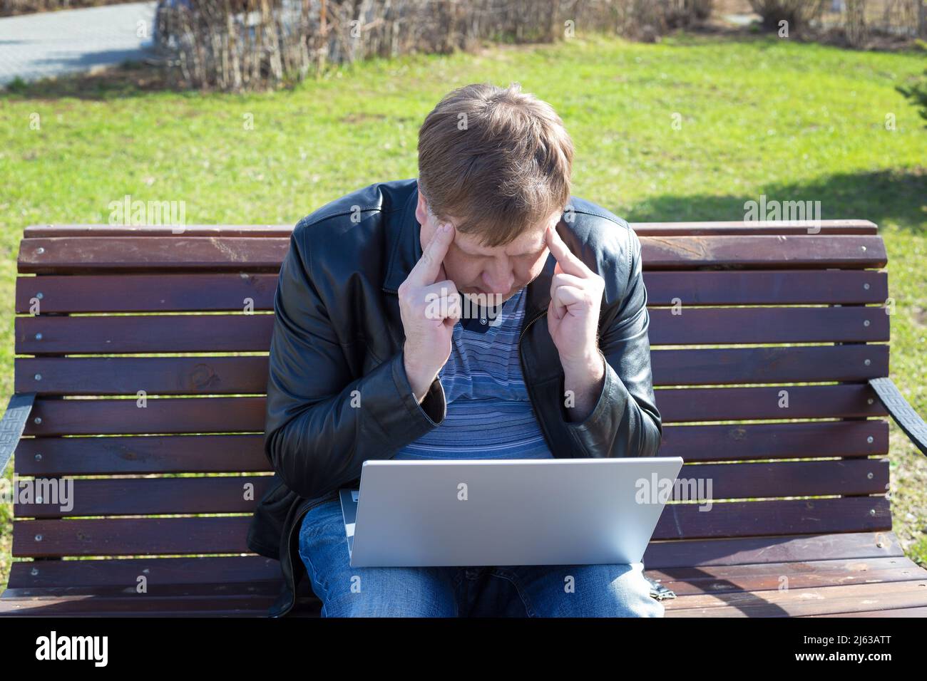 Ein müder, blonder Mann von 40-45 Jahren klammerte sich vor Schmerzen am Kopf, während er an einem Computer arbeitete, während er auf einer Bank saß. Stockfoto