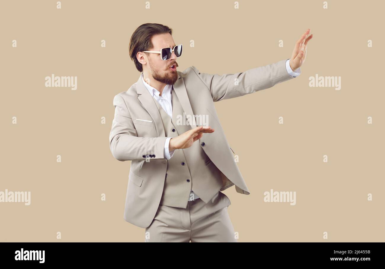 Lustiger junger Mann im klassischen Anzug und coole Sonnenbrillen tanzen auf beigem Studiohintergrund Stockfoto