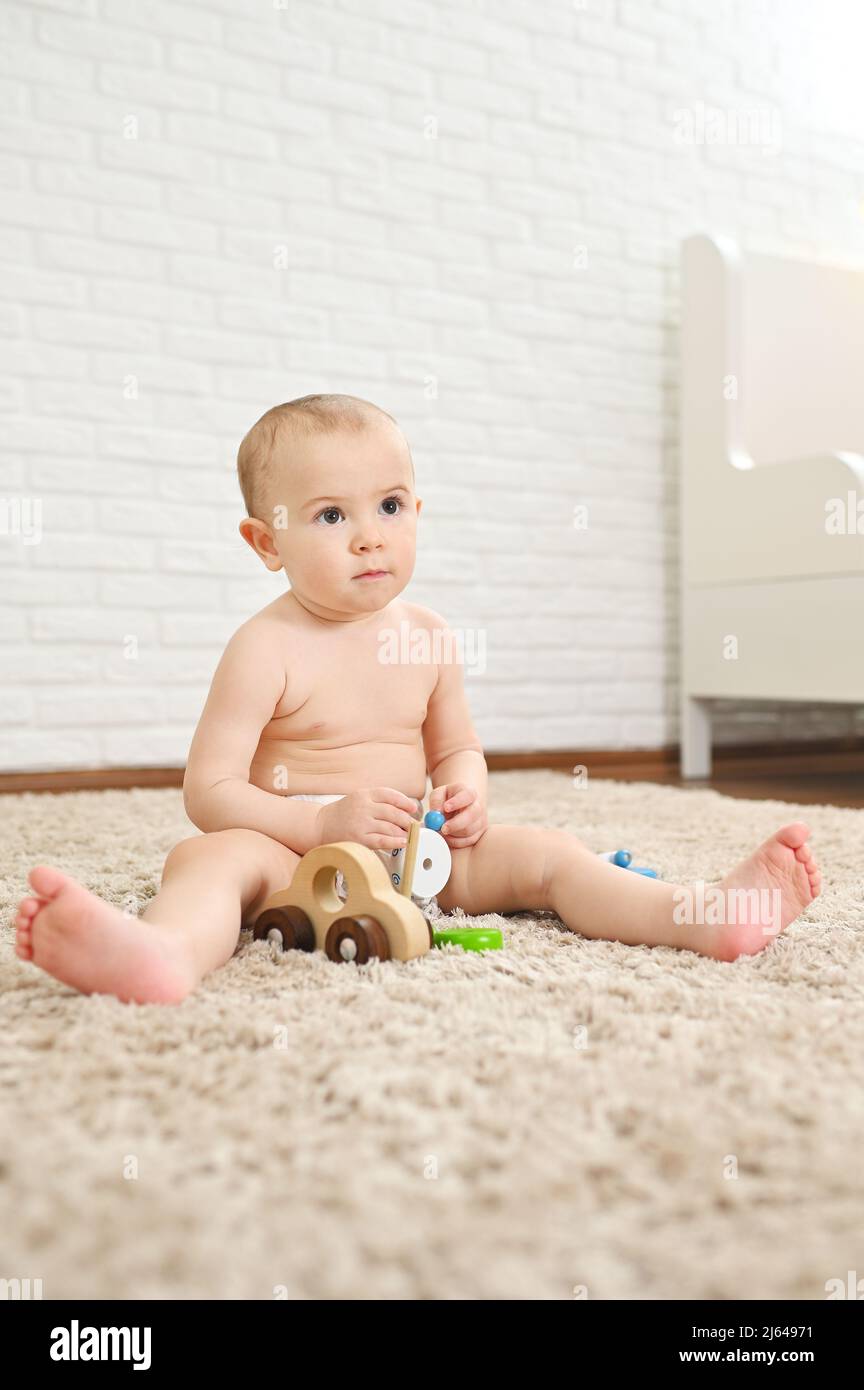 Niedliches Baby sitzt auf einem Teppich in einer Einwegwindel spielt mit Spielzeug gegen eine weiße Ziegelwand. Stockfoto