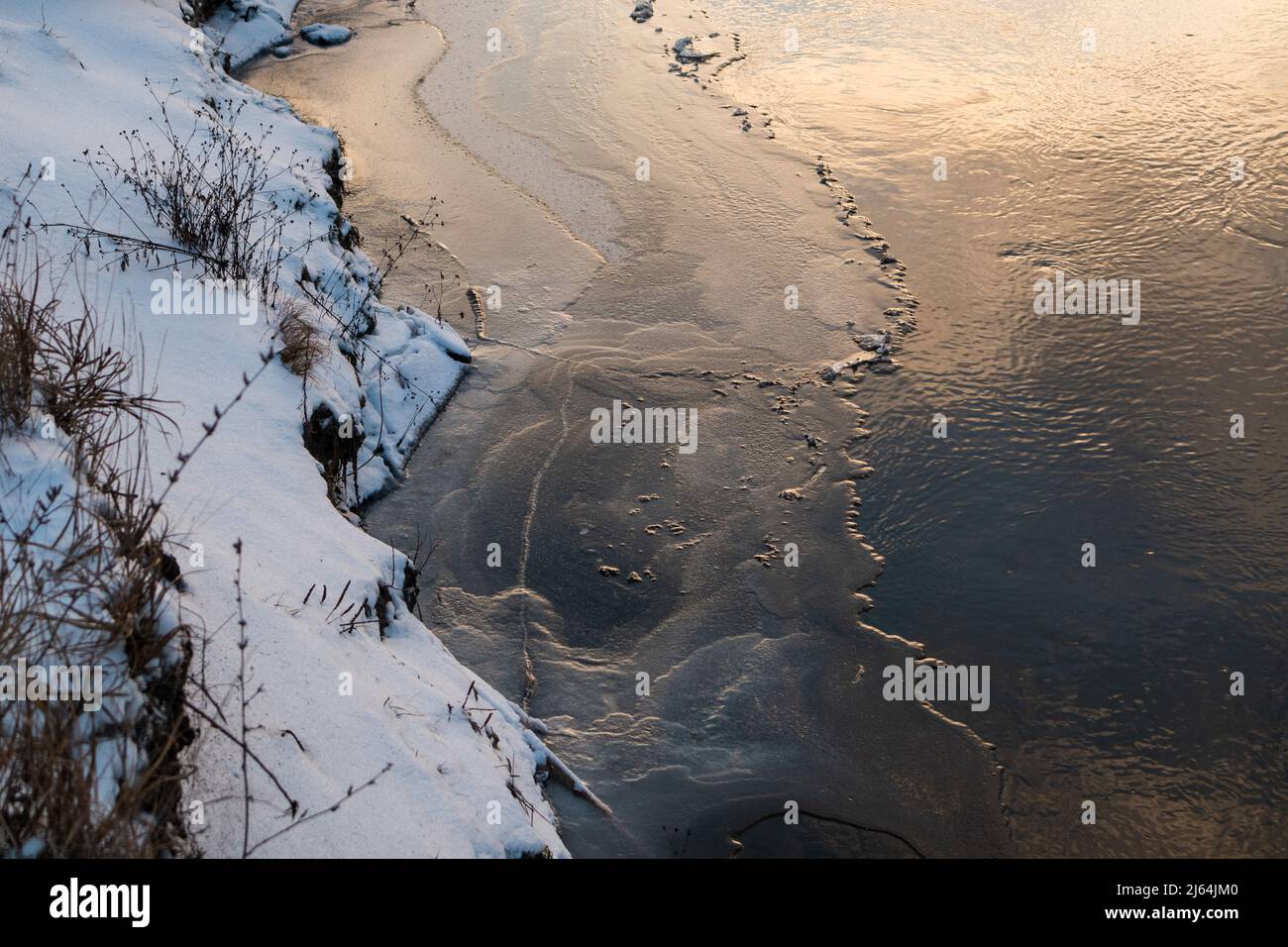 Winteransicht auf kalt verschneiten gefrorenen Flussufer mit trockenem Gras und malerischen Wolken Reflexion. Der Wasserfluss auf dem mit Schnee bedeckten See aus nächster Nähe Stockfoto