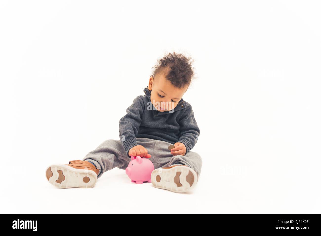 Liebenswert niedlichen Biracial Baby sitzt in einem Studio und setzen Geld in seine rosa Sparschwein über weißem Hintergrund. Einsparungen und zukünftiges Konzept. Hochwertige Fotos Stockfoto