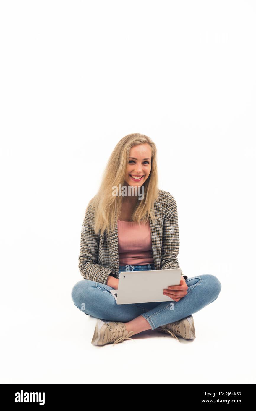 Schöne europäische blonde Mädchen sitzt mit gekreuzten Beinen auf dem Boden lachend und hält einen Laptop voller Schuss Studio Schuss weißen Hintergrund kopieren Raum vertikal . Hochwertige Fotos Stockfoto