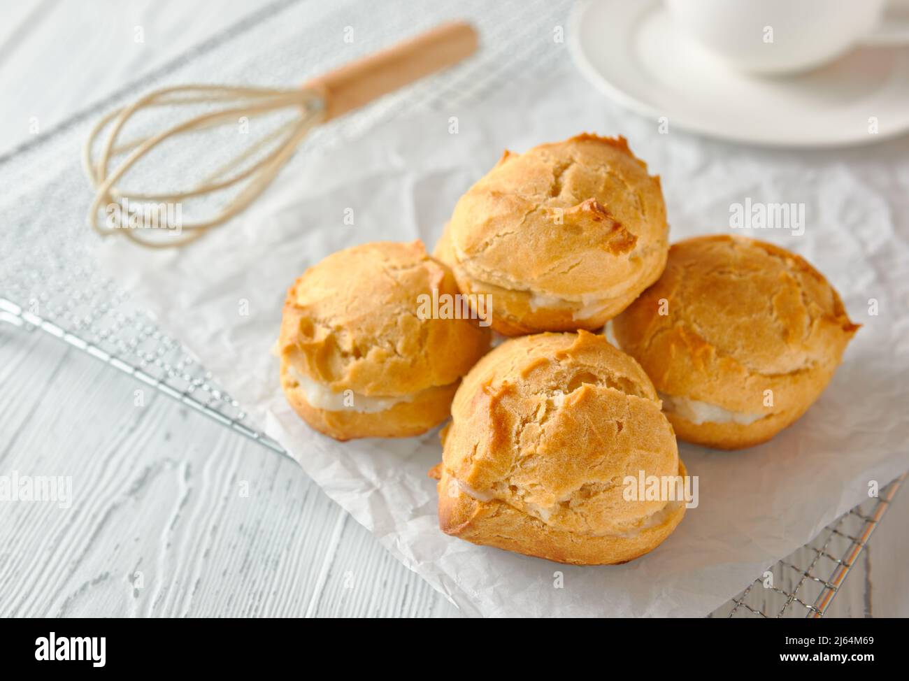 Selbstgemachte Profiterolen oder Eklare auf einem Metallgitter. Daneben befindet sich ein Schneebesen, dahinter steht eine Tasse auf einer Untertasse. Traditionelles französisches Dessert. Weiß Stockfoto