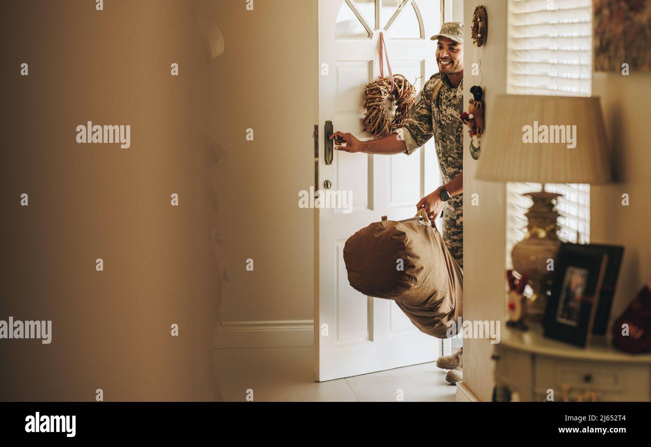 Fröhlicher junger Soldat, der fröhlich lächelt, als er mit seinem Gepäck in sein Haus kommt. Patriotischer amerikanischer Militärangehöriger, der nach Serv eine Überraschungsreise nach Hause macht Stockfoto