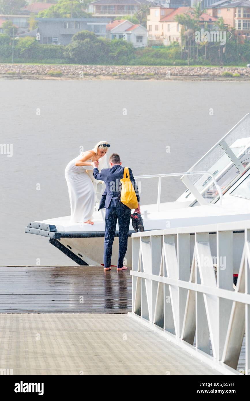 Der gerade verheiratete Kieran hilft seiner neuen Braut Amy, barfuß und im Regen, von einem Wassertaxi in Sydney an Land zu kommen, auf dem Weg zu ihrem Empfang Stockfoto