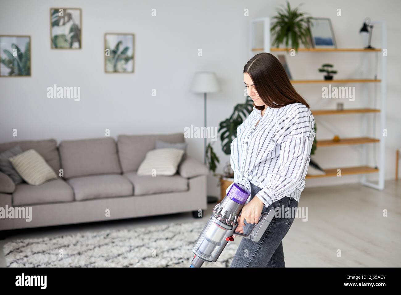 Frau, die den Boden mit einem kabellosen Handstaubsauger im Wohnzimmer zu Hause reinigt. Hausfrau bei der Hausarbeit Stockfoto