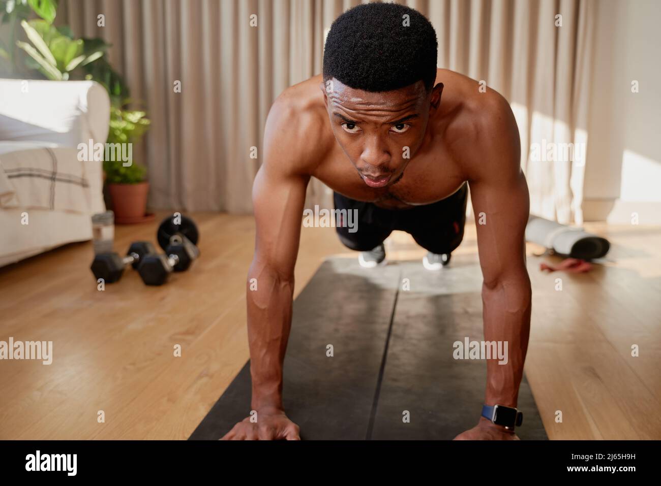 Afroamerikanischer junger Mann mit hoher Planke in seinem warm beleuchteten Wohnzimmer, der zu Hause arbeitet und dabei ist, einen Push-up zu machen Stockfoto