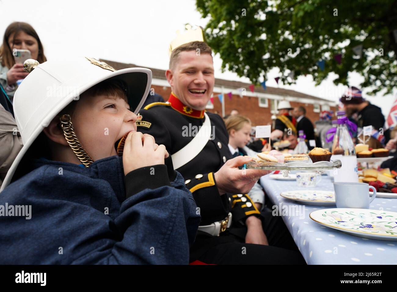 Will Hughes, 7, isst einen Kuchen, während er den Hut von Bandmitglied Aaron Pitman trägt. Die Bewohner von Church Hill in Redditch wurden von Mitgliedern des Royal Marines Band Service überrascht, Die im Juni dieses Jahres ein Medley von Melodien gespielt haben, um die Gemeinde dafür zu würdigen, dass sie sich verstärkt hat, um das Platin-Jubiläum von Königin Elizabeth zu feiern, wobei mehr als 40 große Jubiläumsfeiern geplant waren, um die Nachbarn auf den Straßen der Stadt zusammenzubringen. Bilddatum: Donnerstag, 28. April 2022. Stockfoto