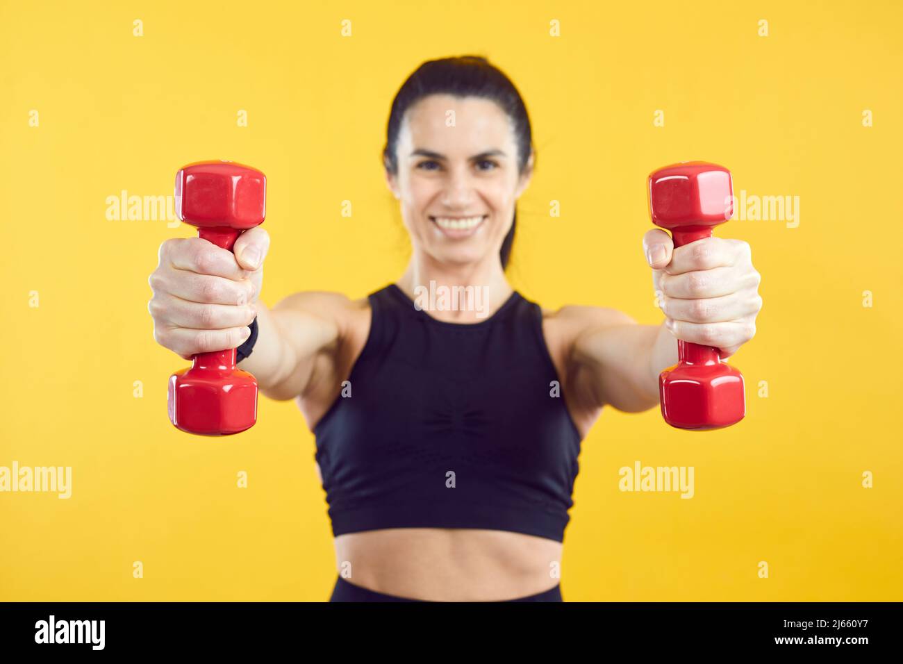 Nahaufnahme von zwei roten Kurzhanteln in den Händen einer sportlichen Frau, isoliert auf lebhaft gelbem Hintergrund. Stockfoto