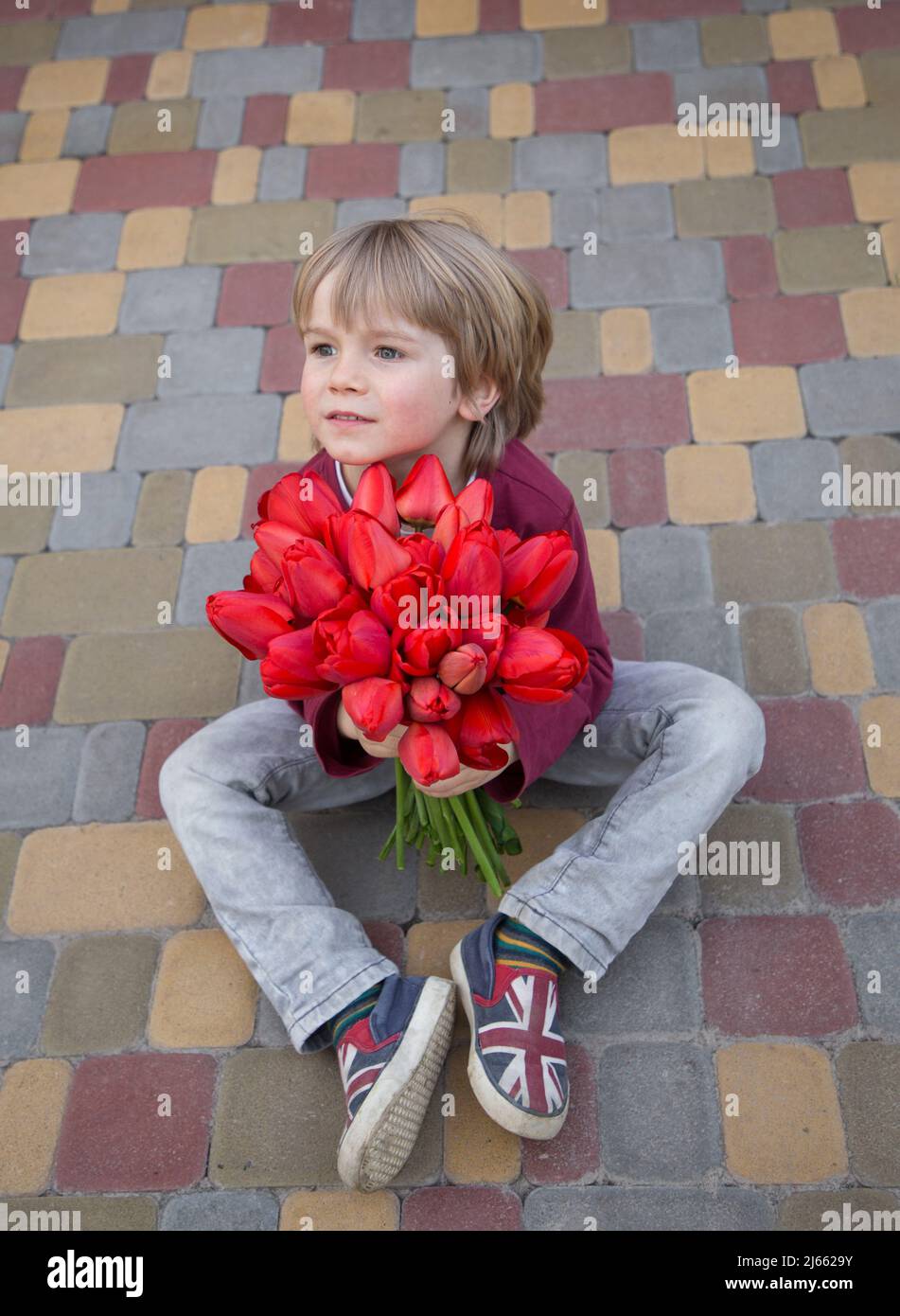 Netter Vorschuljunge sitzt auf einer Pflasterplatte mit einem großen Bouquet aus roten Tulpen. Ein Geschenk mit Liebe zu Mama, Großmutter. Muttertag. Dankbarkeit. Frühlingsmeere Stockfoto