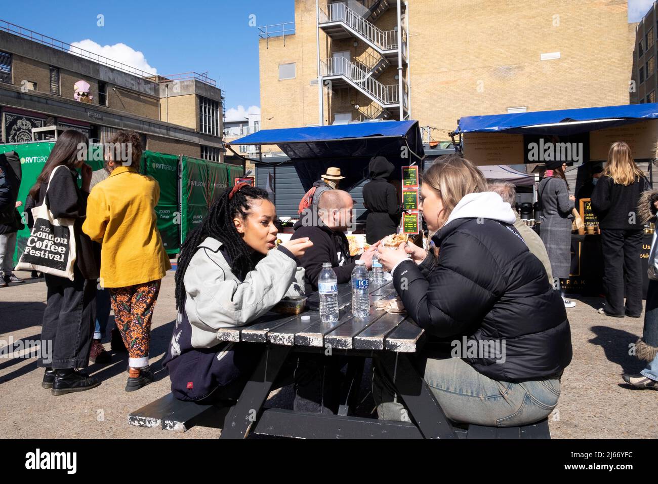 Junge Freundinnen sitzen draußen und unterhalten sich beim Essen von Straßenessen von Stlls in Elys Yard Old Trumans Brewery in der Nähe der Brick Lane East London, Großbritannien, KATHY DEWITT Stockfoto