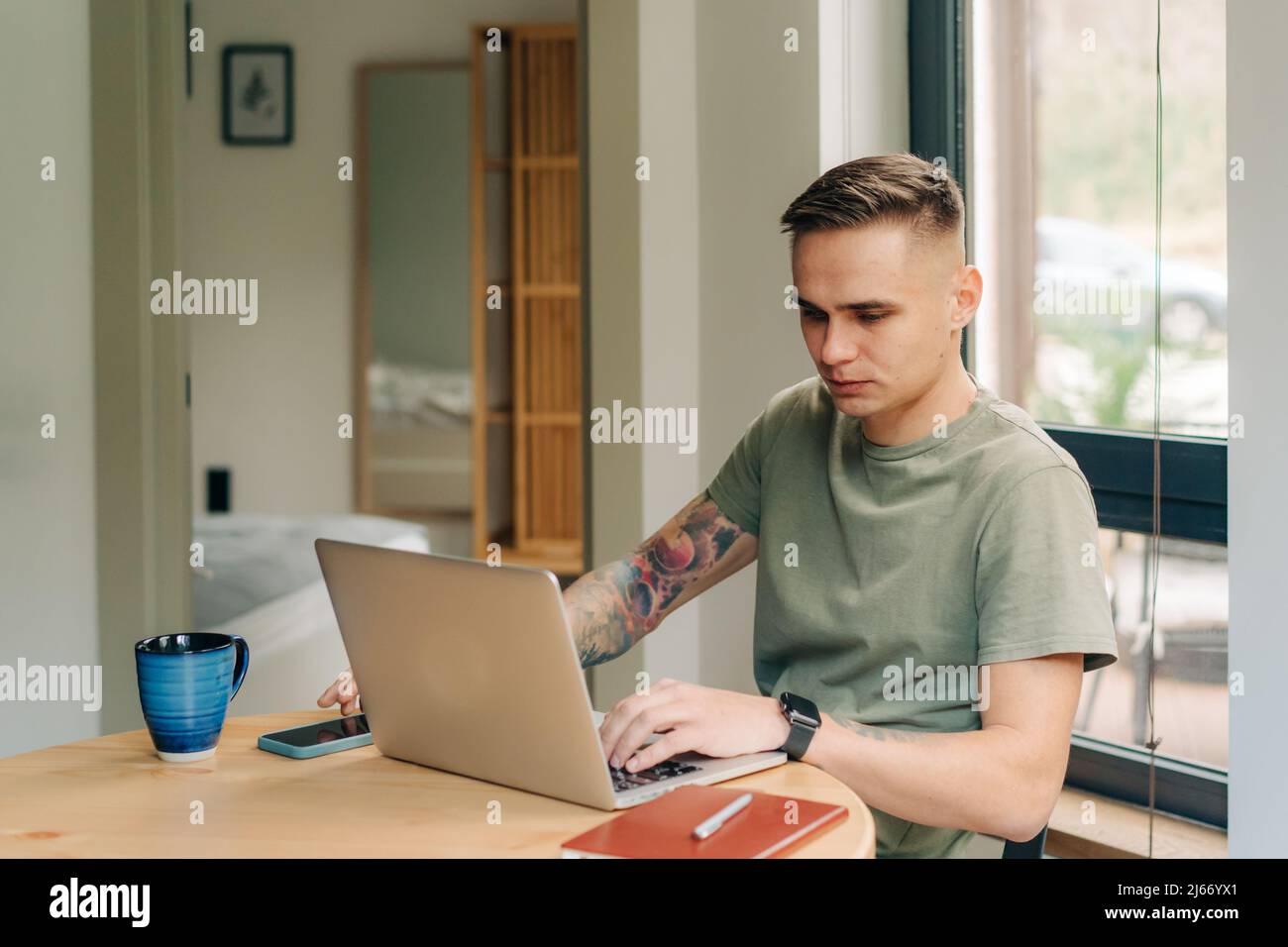 Junger Hipster-Freiberufler, der versucht, sich auf harte Arbeit zu konzentrieren, während er mit einem Laptop am Tisch sitzt. Stockfoto