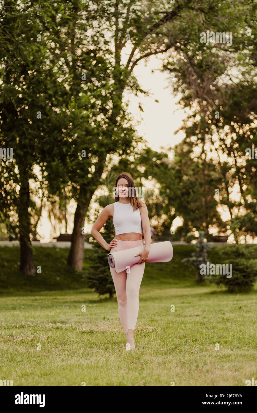 Brunette steht auf Gras im Park. Junge lächelnde Frau in weißem Top und rosa Jogginghose steht auf grünem Rasen und hält rosa Yoga und Stockfoto