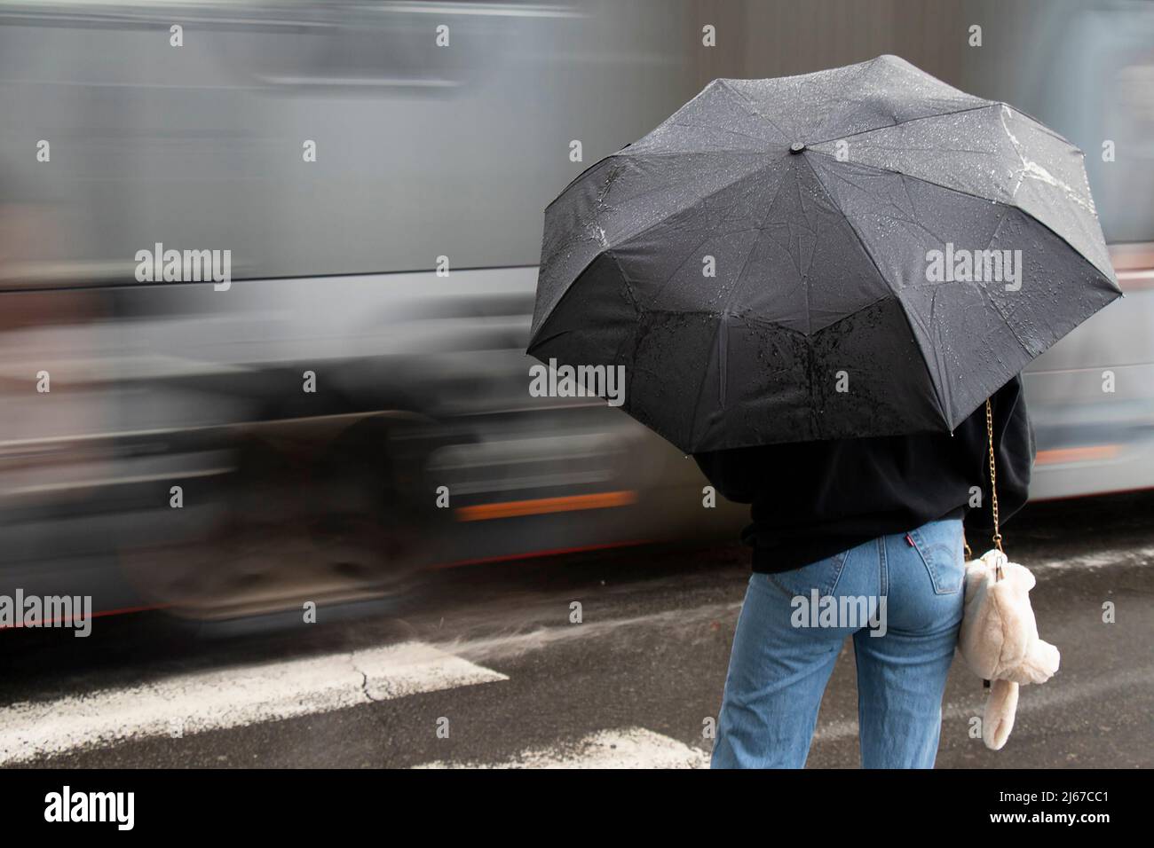 Teenager-Mädchen in Jeans unter schwarzem Regenschirm an einer Kreuzung an einem regnerischen Tag mit Verkehrsunschärfe, Rückansicht Stockfoto
