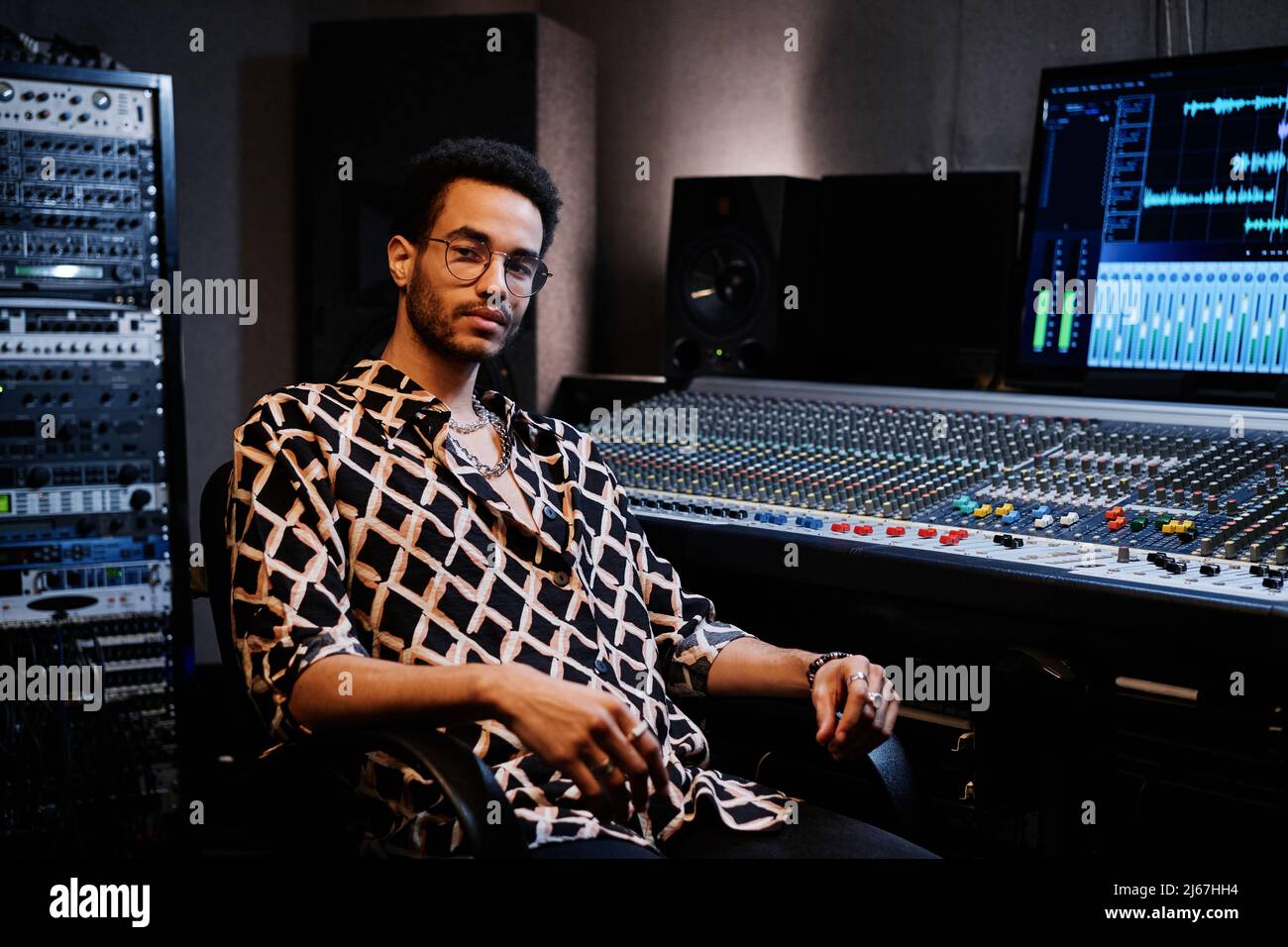 Der junge afroamerikanische Produzent trägt ein modisches Outfit und sitzt im Studio gegen eine Mischkonsole Stockfoto