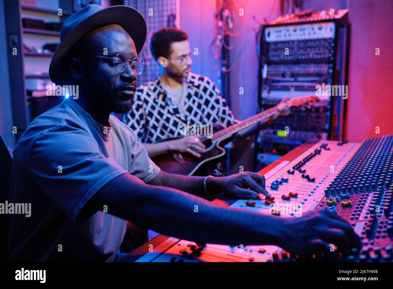 Selektive Fokussierung des stylischen, reifen afroamerikanischen Produzenten, der den Sound auf dem Mixer justiert, während ein junger Musiker Gitarre spielt Stockfoto