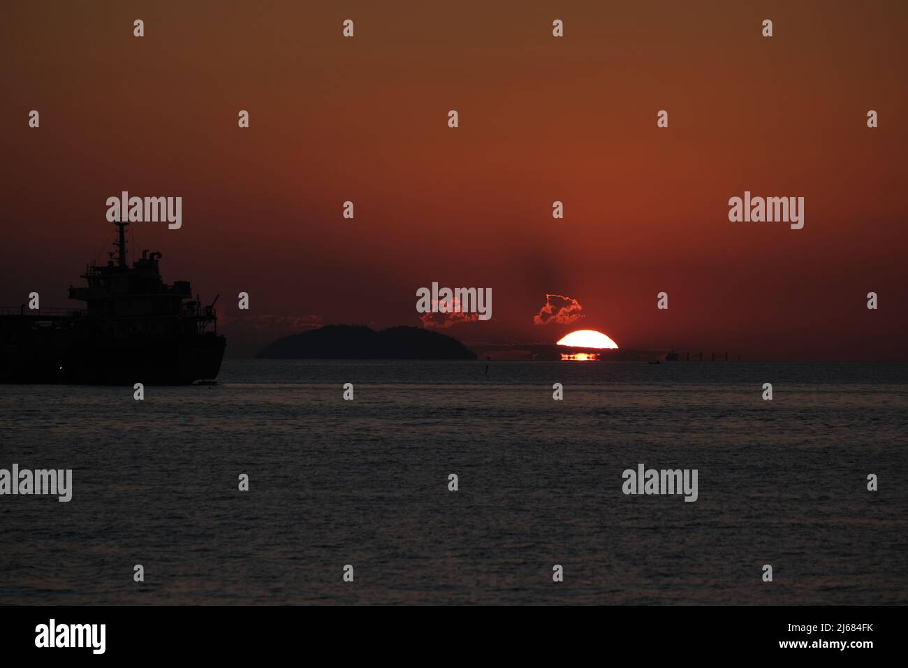 Letzte Sekunden eines Sonnenuntergangs über dem Meer, mit einem großen Schiff und einer entfernten Insel als Silhouetten gesehen Stockfoto