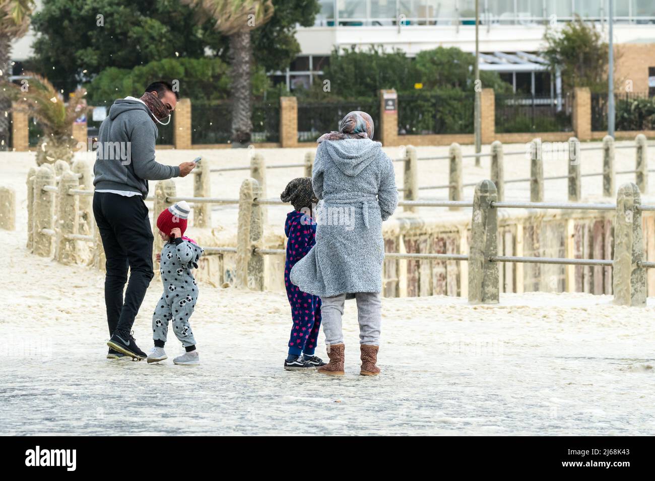 Junge Familie oder Gruppe von Menschen in Winterkleidung, die während der stürmischen Windverhältnisse an der Küste in Kapstadt, Südafrika, in Meeresschaum stehen Stockfoto
