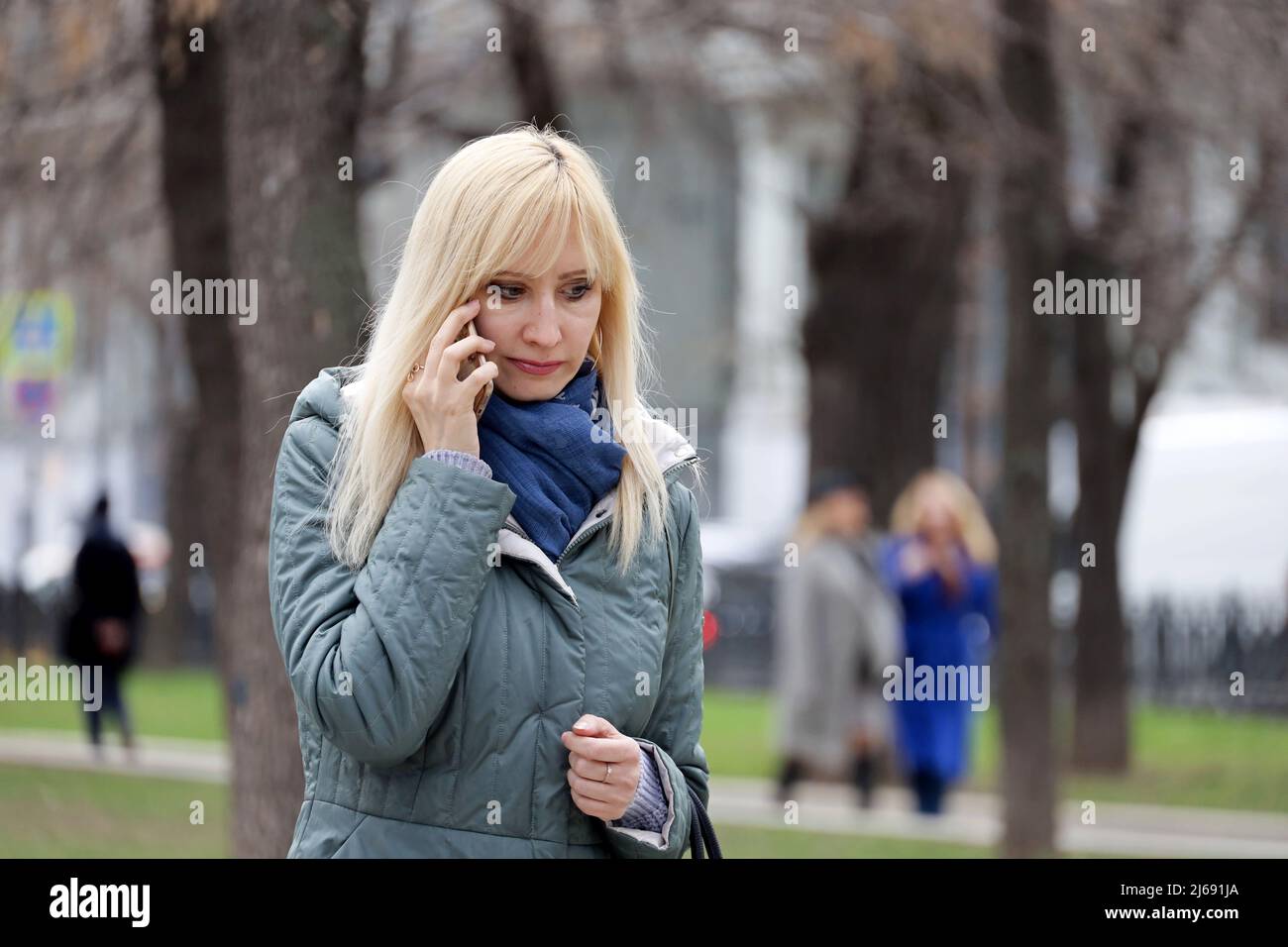 Blonde Frau im Mantel spricht auf dem Handy auf einer Straße in der Frühlingsstadt zu Fuß Stockfoto