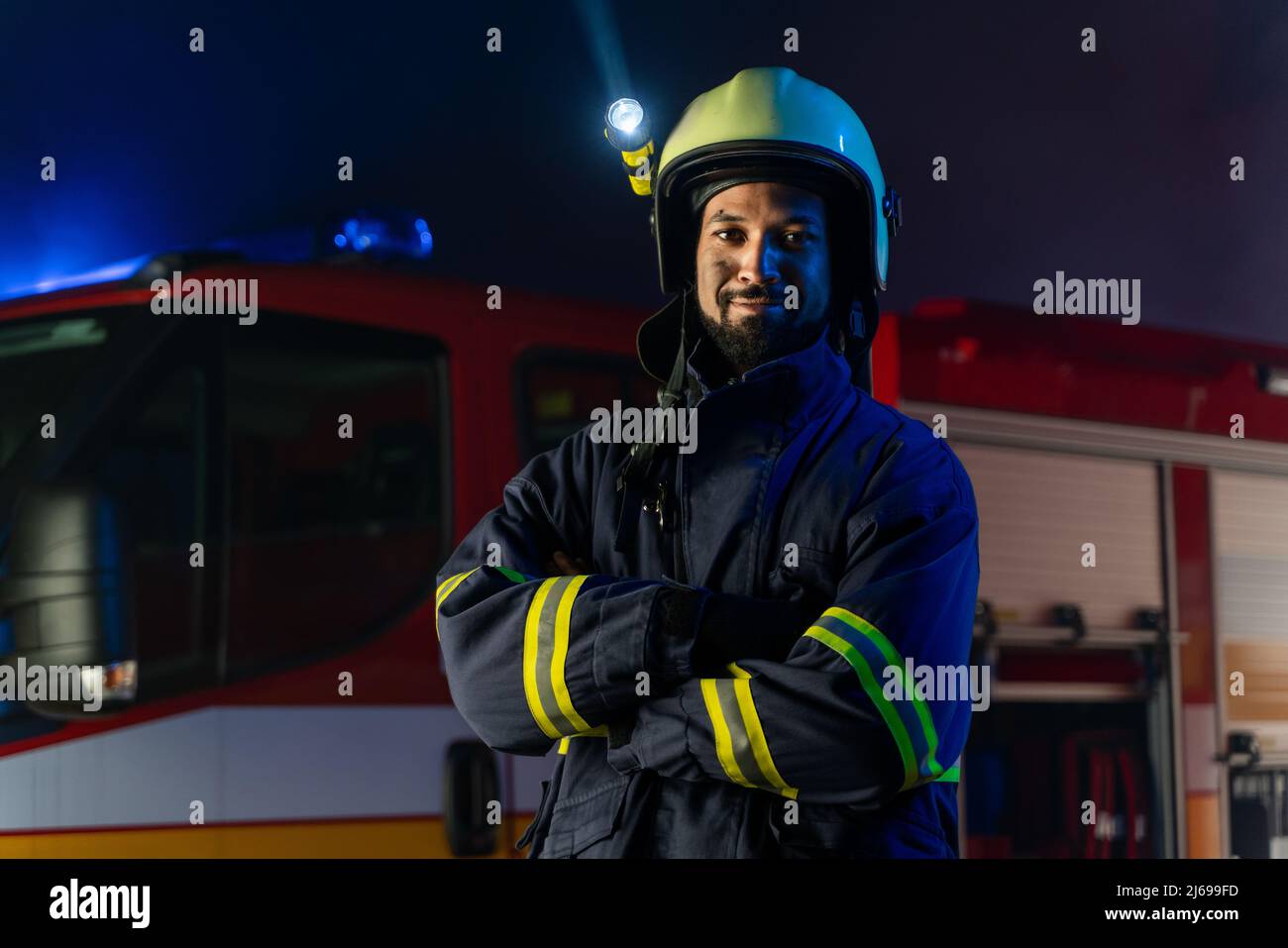 Porträt eines schmutzigen Feuerwehrmanns im Dienst mit einem Feuerwehrauto im Hintergrund in der Nacht, lächelnd. Stockfoto