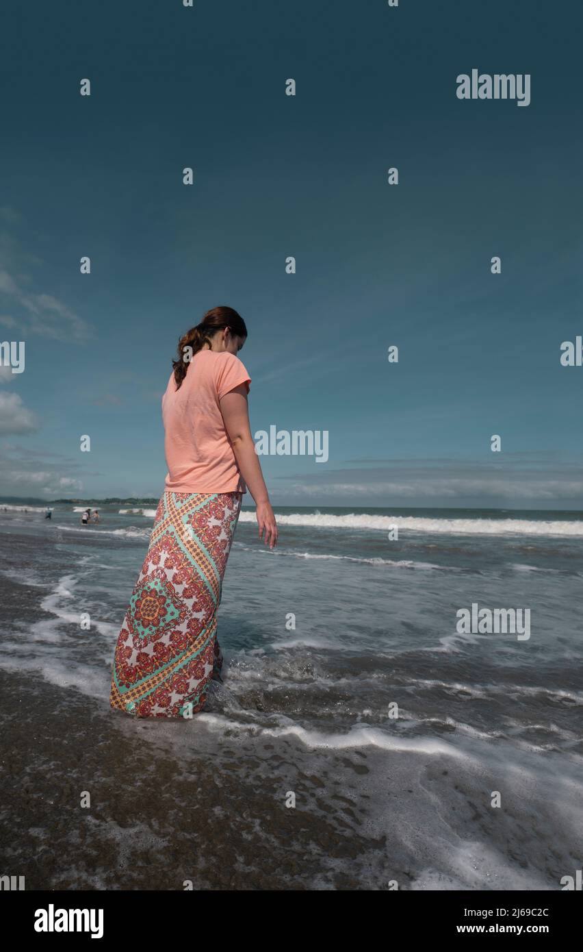 Schöne junge hispanische Frau in einem rosa Kleid von hinten gesehen, die an einem sonnigen Morgen am Strand mit dem Meer im Hintergrund spazieren ging Stockfoto