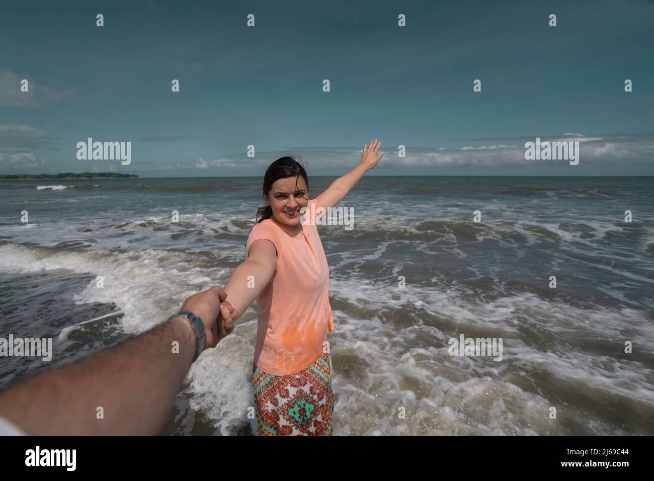 Schöne, glückliche junge hispanische Frau, die die Hand ihres Partners am Strand hält und während eines sonnigen Mornis ein rosa Kleid mit dem Meer im Hintergrund trägt Stockfoto