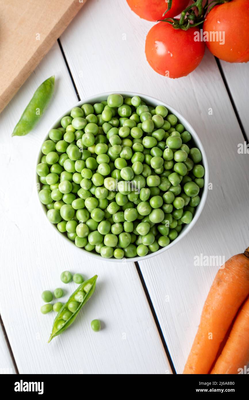 Frische, süße Erbsenschoten. Grüne Erbsen in weißer Schale mit frischen Schoten auf dem weißen Holztisch. Draufsicht. Stockfoto