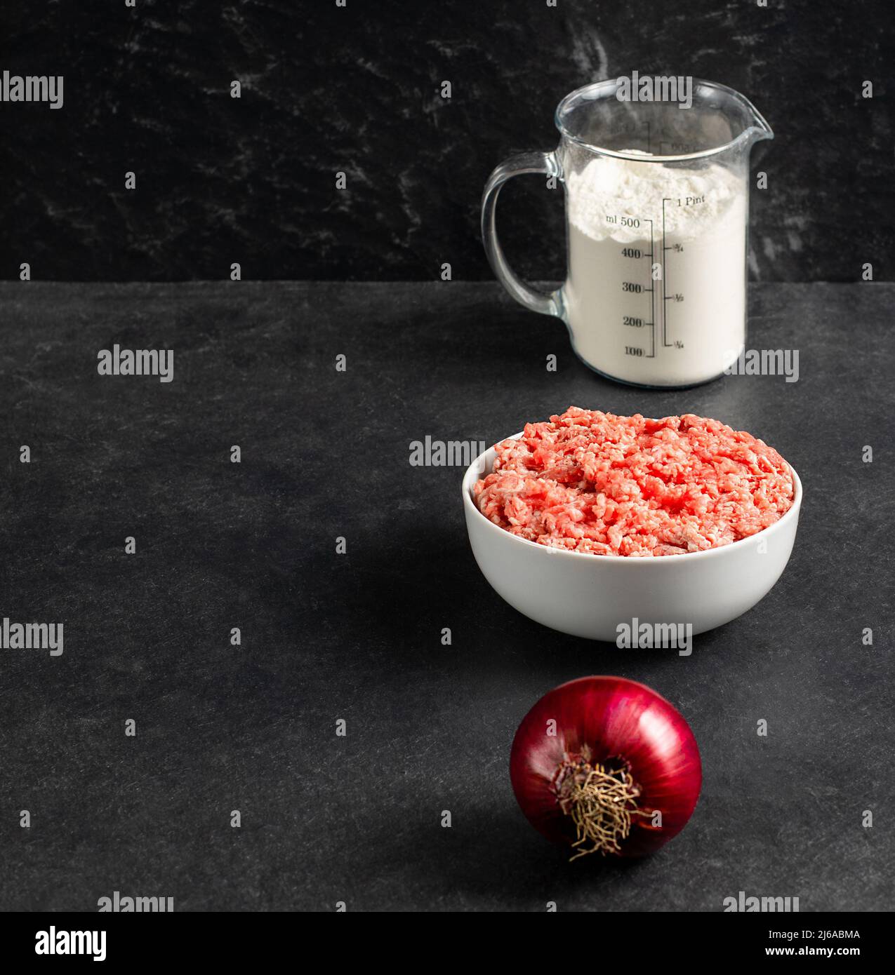 Teller mit frisch gemahlenem Rindfleisch, roter Zwiebel, Messbecher mit Mehl auf grauem schwarzem Hintergrund, Platz für Text, Stockfoto Stockfoto
