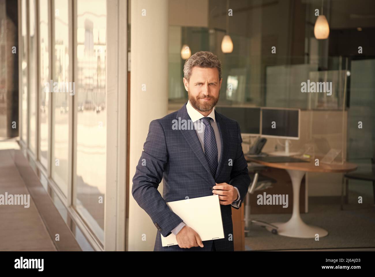 Ein reifer, lächelnder Geschäftsmann in einem Geschäftsanzug hält einen drahtlosen Laptop vor dem Büro, dem Geschäft Stockfoto