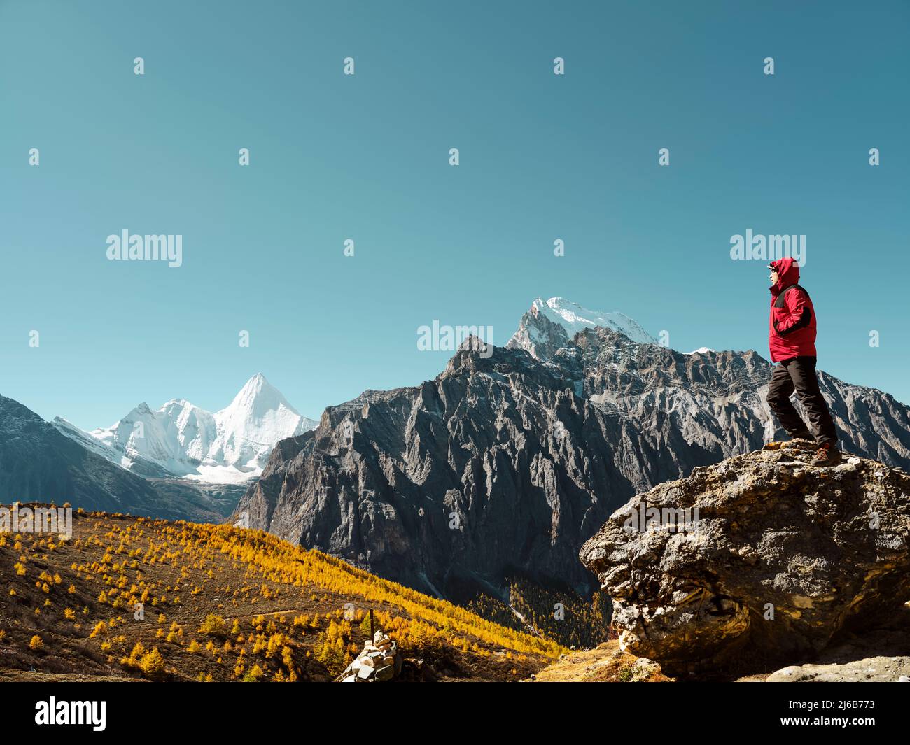 ein asiatischer Mann, der auf einem Felsen steht und den Berg Yangmaiyong (oder auf Tibetisch Jampayang) in der Ferne in Yading, Kreis Daocheng, Sichuan, anschaut Stockfoto