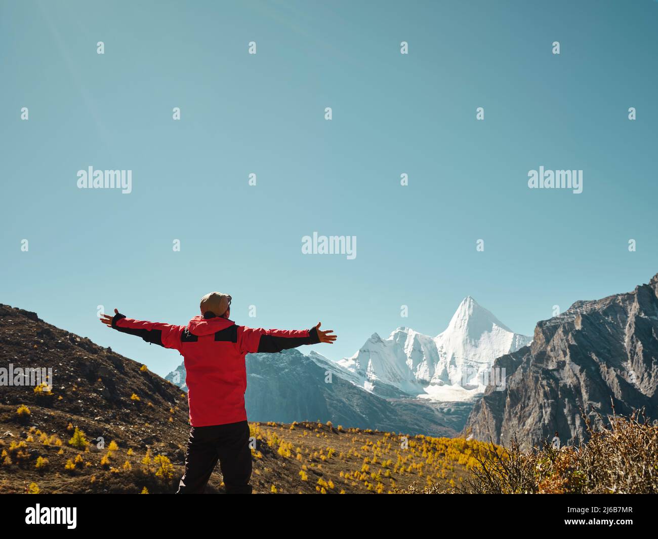 Rückansicht eines asiatischen Mannes, der auf einem nahe gelegenen Berg steht und den Berg Yangmaiyong (oder Jampayang auf Tibetisch) mit offenen Armen in Yading, Daocheng, anschaut Stockfoto