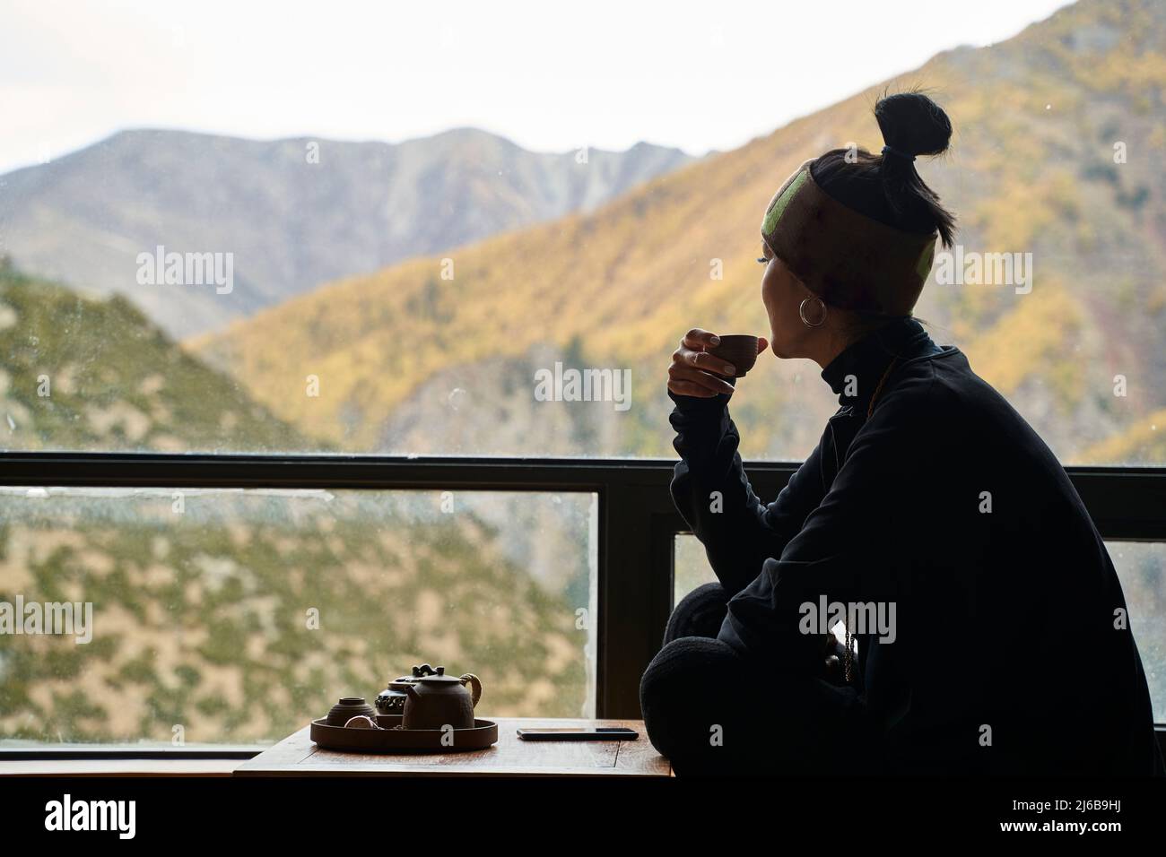 asiatische Frau, die auf dem Balkon sitzt und Tee trinkt und die Berglandschaft mit herbstlicher Vegetation genießt Stockfoto