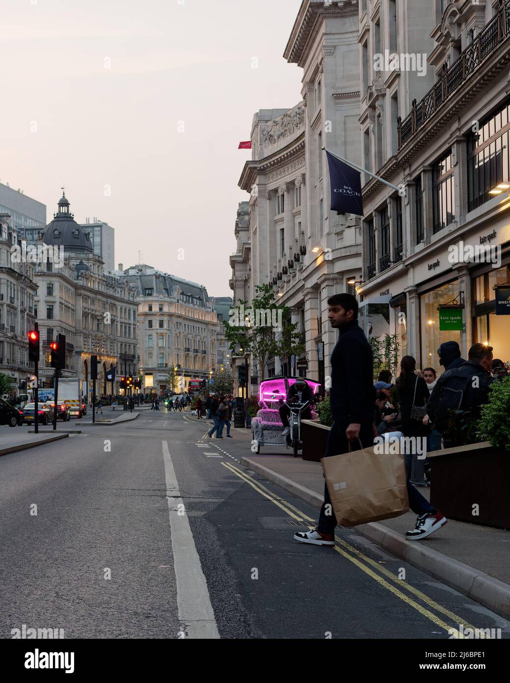 London, Greater London, England, 23 2022. April: Ein Mann mit einfacher Einkaufstasche überquert abends die Regent Street mit einer rosa Rickshaw aka Pedicab aka Stockfoto
