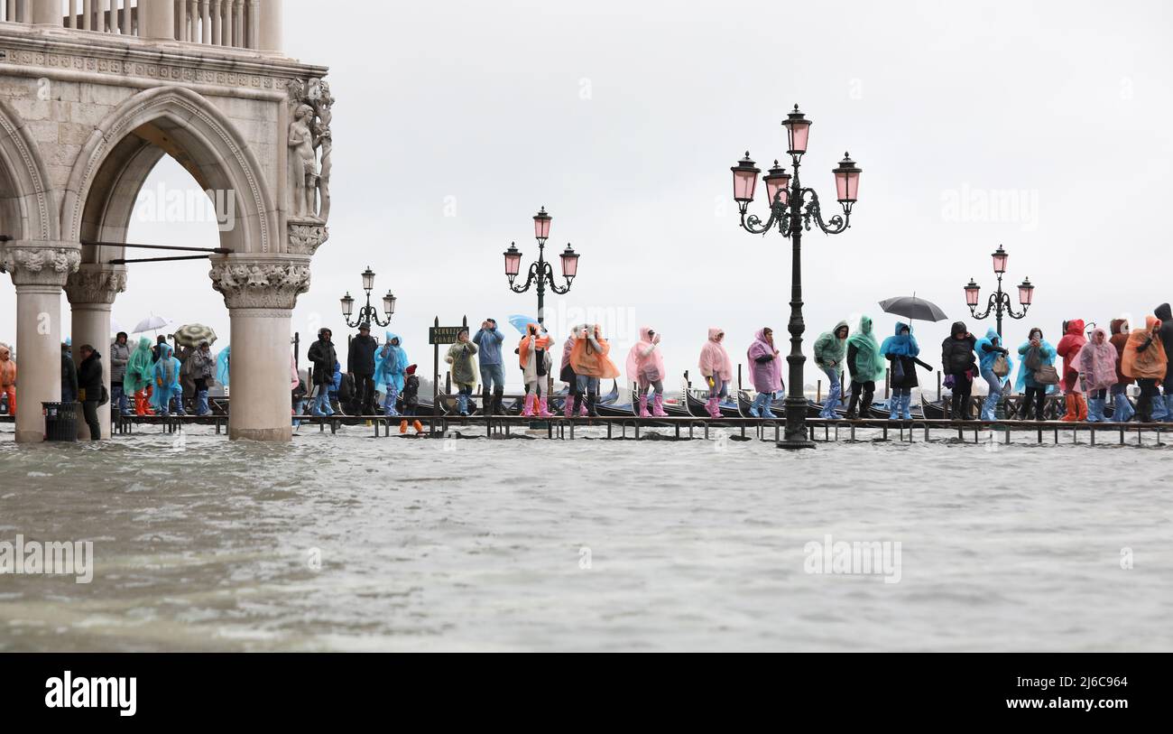 Menschen mit Regenkleidung gehen bei Flut auf dem erhöhten Weg auf dem unter Wasser liegenden Markusplatz in Venedig in Italien in Europa Stockfoto