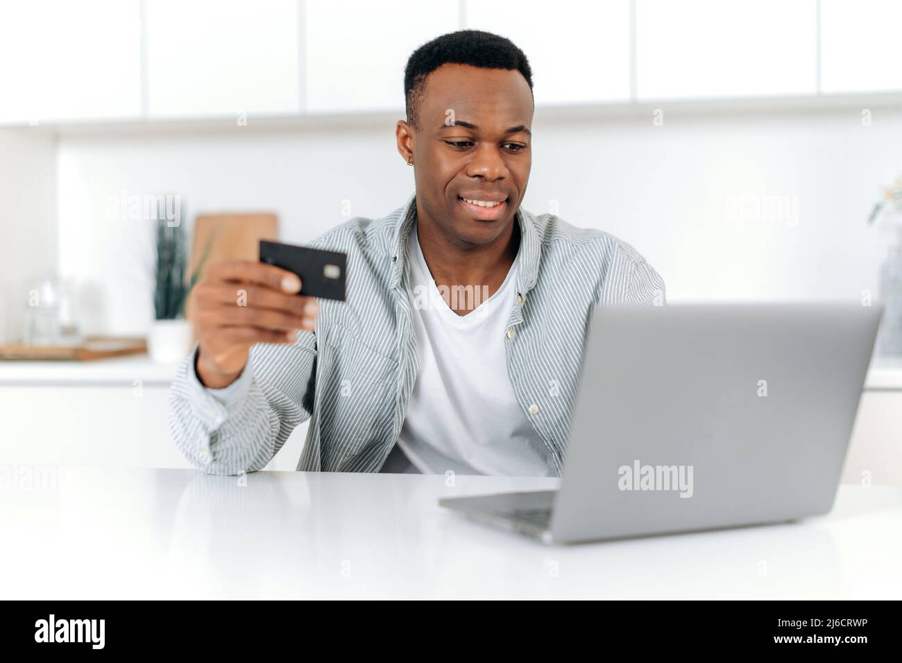 Glücklicher afroamerikanischer Mann, der zu Hause sitzt, in lässiger Kleidung gekleidet, Laptop und Kreditkarte zum Online-Shopping verwendet, Daten für die Zahlung eingibt, Online-Transaktion, Lieferung nach Hause bestellen, lächelnd Stockfoto