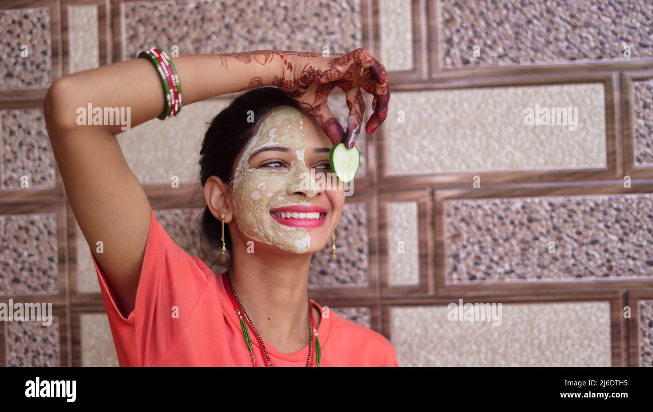Eine Frau mit Gesichtsmaske und frischer Gurke auf den Augen, die sich zu Hause entspannt. Nahaufnahme Porträt eines lächelnden Mädchen in Ton-Maske. Hausgemachte Multani mitti Gesicht Pac Stockfoto