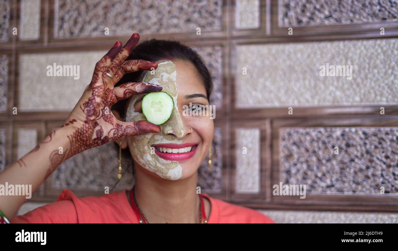 Eine Frau mit Gesichtsmaske und frischer Gurke auf den Augen, die sich zu Hause entspannt. Nahaufnahme Porträt eines lächelnden Mädchen in Ton-Maske. Hausgemachte Multani mitti Gesicht Pac Stockfoto