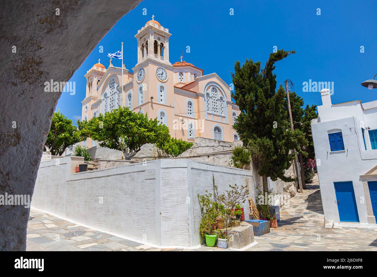 Tinos, Griechenland - 1. Juli 2021: Traditionelle Kirche mit rosafarbenem Mauerwerk, großer Turmuhr und einer im Wind winkenden griechischen Flagge. Blick durch einen Torbogen, bergauf Stockfoto