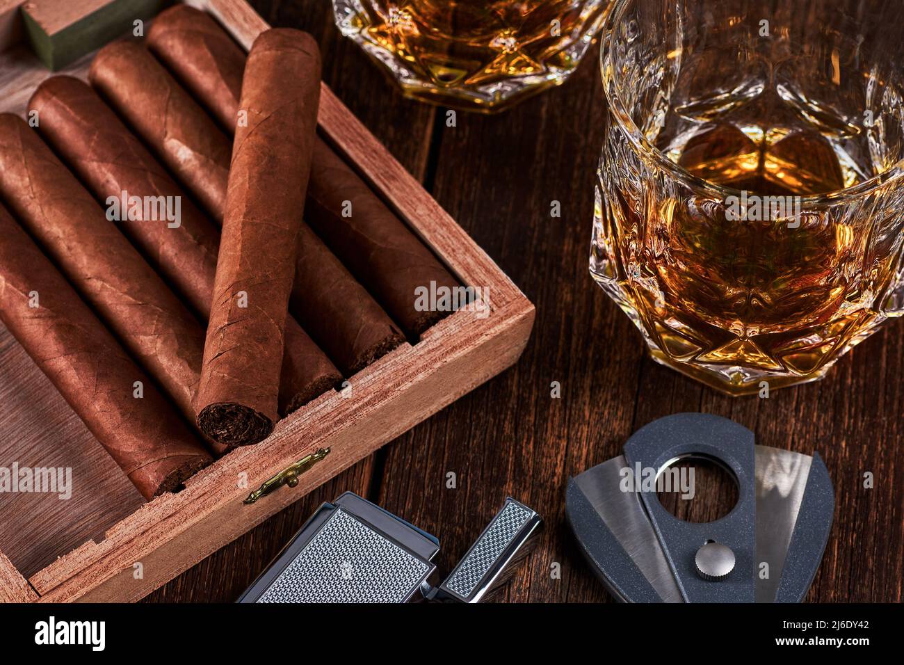 Box mit kubanischen Zigarren, Feuerzeug und Cutter auf alter Holztischplatte. Zwei Gläser Whiskey oder Alkohol auf dem Hintergrund. Stockfoto
