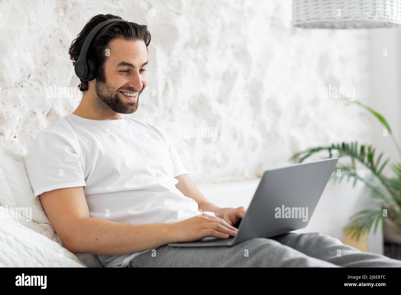 Glücklicher junger Mann, der auf dem Bett saß und den Computer benutzte Stockfoto